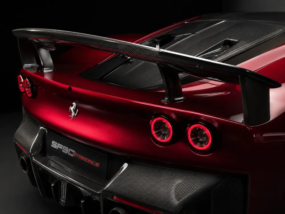 Close-up studio image of a Ferrari rear end fitted with a sleek carbon fiber wing, showing visible carbon weave, aerodynamic curves, dramatic lighting, and a premium performance-car atmosphere