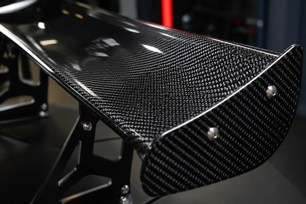 Detailed macro image of a carbon fiber wing surface with glossy clear coat, tight carbon weave pattern, mounting brackets, and workshop lighting for a luxury sports car part