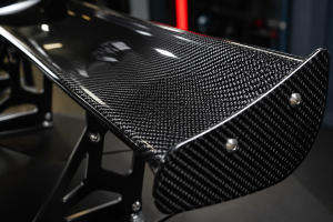 Detailed macro image of a carbon fiber wing surface with glossy clear coat, tight carbon weave pattern, mounting brackets, and workshop lighting for a luxury sports car part